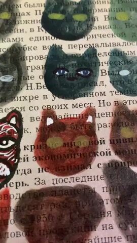 The media file shows a close-up view of a piece of artwork being created. The artwork is composed of several painted cat faces, each with unique colors and expressions, applied over a background of text from a book. The text is in Russian.

The primary subject is the hand of a person, holding a fine-tipped tool, likely a pen or a fine brush, and meticulously adding detail to one of the painted cat faces. The hand is seen from a slightly elevated angle, with fingers obscuring parts of the artwork.

The scene is an intimate, artistic process. The focus is on the act of drawing or painting the details, specifically the whiskers and mouth of a dark green cat. The cat has large, expressive blue eyes. Other cat faces in varying shades of black, brown, and red are visible around the central cat, some with yellow or green eyes, and some with less defined features. The texture of the paint and the paper background is visible.

Notable details include the unfinished nature of the artwork, with the artist actively working on it. The lighting appears to be indoor, and the overall mood is one of concentration and creative engagement. The Russian text on the background suggests a potential connection to Russian literature or culture, although the specific content is not legible enough to decipher fully. The location context provided is Sergeevo, Russia.