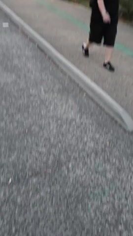 The video captures a person in a black shirt and pants walking on a paved path. The path appears to be part of a road or walkway, with a curb separating it from a lane of traffic where a car is visible. The environment suggests an outdoor setting, possibly urban or suburban, with trees and streetlights. The sky in the background shows the fading light of dusk, with a mix of blue and pink hues. The person is captured from a low angle, and their movement suggests they are walking away from the camera.