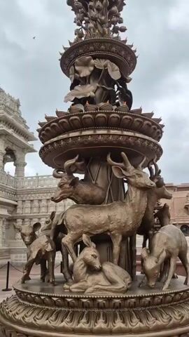 This is a vertical, medium shot video showing a large, ornate bronze fountain in the foreground. The fountain has multiple tiers and is decorated with lotus flower motifs. Surrounding the base of the fountain are several bronze statues of deer, some standing and some lying down. In the background, to the left, is a large, intricately carved white stone building that appears to be a temple. The building has multiple spires and decorative carvings. The sky above is overcast. In the middle ground, a person wearing a blue and white checkered shirt and dark pants walks across a paved courtyard. Further to the left, a person in an orange shirt and dark pants is kneeling on the ground. There are black metal posts with ropes or chains in the foreground, separating the viewer from the courtyard. Two dark trash cans are visible in the courtyard. The video pans slightly to the left, revealing more of the temple structure.