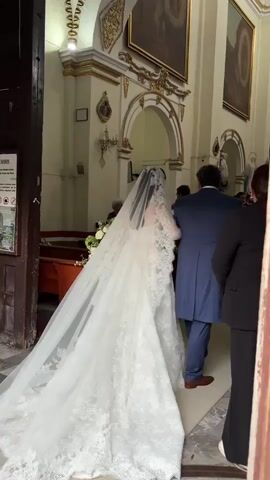 Inside a church, a bride in a white lace wedding gown with a long veil is escorted down the aisle by a man in a dark blue suit. The bride’s veil is lace-edged and trails behind her. The man holds the bride’s arm to steady her. A woman in a black jacket walks beside them. On the left side of the aisle is a large arrangement of white flowers. Several people are seated in pews, while others stand and watch the bride pass. The church interior is ornate with gilded accents and arched alcoves. A painting hangs on the wall. A woman kneels down to adjust the bride’s veil as she passes.