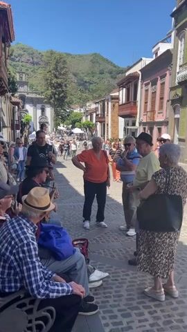 The video shows a street scene in a town with traditional architecture. In the foreground, several people are gathered, some sitting on a bench and others standing. A man is playing an instrument that appears to be a guitar. In the background, more people are walking and there are buildings with balconies. The sky is blue and there are mountains in the distance. The overall impression is of a lively public space.