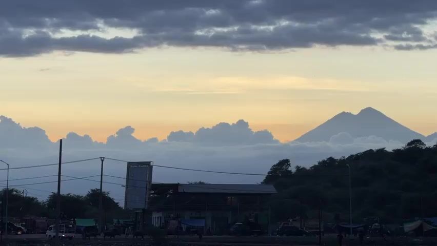 The video showcases a picturesque scene with a large mountain towering over a bustling coastal town. The mountain is partially obscured by clouds, giving it a mystical and ethereal appearance. The town is bustling with activity, with vehicles and people moving around. The sky is adorned with streaks of colors, indicating either sunrise or sunset. The overall atmosphere is serene and captivating, capturing the beauty of nature and human activity coexisting in harmony.