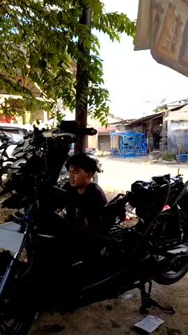 The video shows an outdoor motorcycle repair shop with two men working on vehicles. The first man is young, wearing a black shirt, and is sitting on a disassembled black motorcycle. The second man is older, wearing a dirty brown shirt and a baseball cap. He is sitting on a wooden structure nearby. Other motorcycles are parked in the background, along with a white car and various tools. The shop appears to be located near a river or canal, visible in the background. The environment is shaded by trees and looks like a rural or semi-rural area.