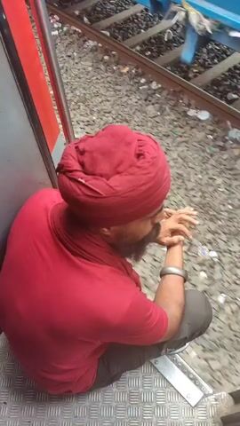 A man wearing a red turban and a red shirt is sitting on the steps of a train car, facing away from the camera. He has a beard and is wearing a silver bracelet. He is holding his hands clasped in front of his face and appears to be looking down. The train car is red and grey. The ground around the tracks is covered in gravel and debris. The train tracks are visible in the background.