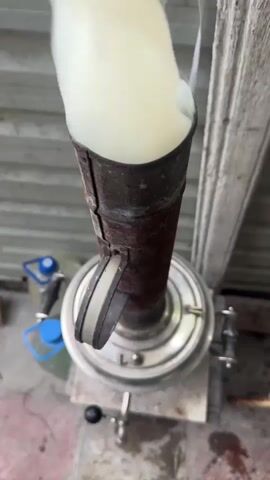 A close-up shot shows a metal tube smoking heavily, with white vapor billowing upwards. The tube appears to be part of a samovar, a traditional Russian teapot. The samovar is made of metal and has handles. In the background, there is a blurry view of a wooden structure and some plastic containers. The lighting suggests an outdoor setting. The video captures the steam escaping from the top of the device.