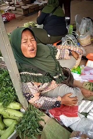 The video shows a woman wearing a green hijab and a patterned top sitting in a market. She is leaning against a pole and appears to be speaking or crying. Around her are piles of vegetables, including green leafy vegetables and what look like cucumbers or gourds. There are also baskets, plastic bags, and a scale. In the background, other people are visible, also in a market setting, with motorcycles and stalls. One person is wearing a brown top and a hijab, and another person is wearing a green hijab. The ground is dirt.