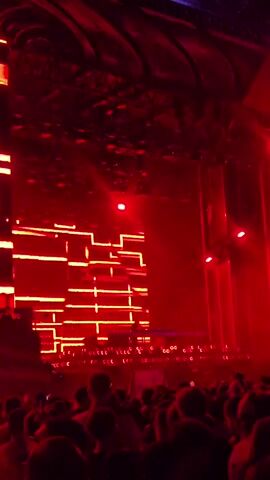 The video captures a concert or DJ set taking place in a dimly lit venue. Red lights illuminate the scene, creating a vibrant and energetic atmosphere. A large LED screen displays abstract, glowing red patterns, forming a backdrop for the stage. In the center of the stage, a DJ is visible behind a console, presumably controlling the music and lights. A crowd of people is gathered in front of the stage, facing the DJ and the visual display, with some holding up their phones to record the event. The ceiling of the venue appears to be ornate and textured, further enhancing the visual spectacle. The overall impression is one of a lively and immersive entertainment experience.