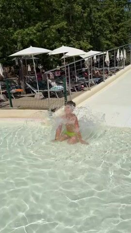 A boy in lime green swim trunks with black trim runs and plays in the shallow end of a water park. He is at the bottom of a large, white water slide and playfully splashes in the water. He looks at the camera, smiles, and sticks out his tongue before turning and running towards a different part of the pool. Other people are visible in the background, including children on the water slide and adults lounging on chairs under umbrellas. The environment appears to be a sunny outdoor water park with lush green trees in the background.