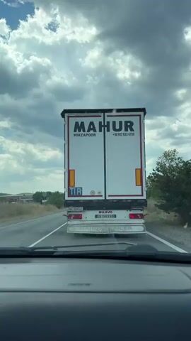 The video shows the rear of a white semi-trailer truck with "MAHUR MIRZAPOOR" written on it. There are speed limit stickers on the back of the trailer, one indicating 70 and another 80. The truck has an Iranian license plate that reads "91AR990 IR". The video appears to be filmed from inside another vehicle, following the truck on a road with dry vegetation on the sides and a cloudy sky.