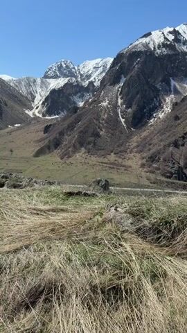 The video shows a mountain landscape on a clear day. In the foreground there is dry, brown grass and some green patches. Several gray rocks are scattered about. A road cuts across a valley in the middle ground, lined by green meadows on both sides. In the background, there are mountains, some of which are snow-capped. The sky is a clear, bright blue. The camera slowly moves slightly forward, changing the composition of the foreground.