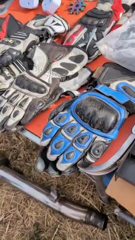 The video shows a close-up, overhead view of a collection of used motorcycle gloves laid out on an orange surface, possibly a table or tarp. The gloves are made of leather and synthetic materials in various colors, including black, white, blue, red, and orange. Some gloves have protective padding, such as carbon fiber accents on the knuckles and fingers. In the background, there are other items that appear to be electronic accessories and possibly some car parts. The ground beneath the table appears to be dry grass or straw, suggesting an outdoor setting like a market or yard sale. The lighting is bright, indicating daytime. No people are visible in the frame.