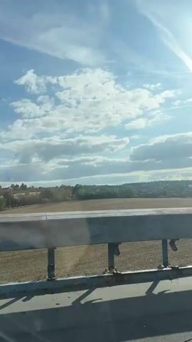 The video shows a view from inside a car driving on a highway. In the foreground, the car's dashboard and windshield are visible. Outside, the highway stretches ahead, with other cars and a white truck with a green logo on the back. The truck has the text "foodora.eu" on its back. The sky is cloudy with patches of blue. On either side of the highway, there are trees and fields. The camera is pointed forward, capturing the road and the landscape. The video appears to be shot in daylight.