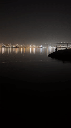 Figure stands on rocks near illuminated bridge in predawn Coronado