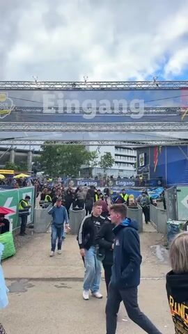 The video shows an outdoor scene at the entrance to an event. Numerous people, predominantly young adults in casual attire, are walking on a dirt path towards and through a series of turnstiles and security checkpoints. Several staff members, distinguished by bright yellow or green safety vests, are present near the entrance points. Overhead, a large banner supported by a metal truss structure displays the word "Eingang" (German for "Entrance") in prominent white letters, along with the "Rock am Ring" event logo on the left. In the background, modern buildings are visible under an overcast sky. To the right of the entrance, a large blue structure with yellow lightning bolt graphics is present. A green tent is partially visible on the far left. The crowd is moving steadily into the event area.