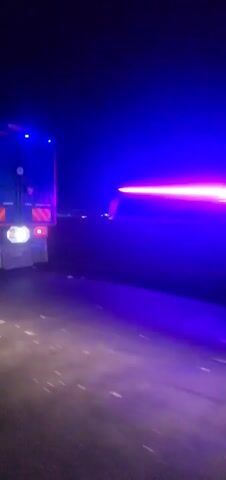 The video, recorded at night, appears to be taken near a roadside, possibly a highway or freeway. Several vehicles are visible, including a large truck with blue flashing lights and an ambulance, also with flashing lights. People are seen moving around near the vehicles. The environment is dark, with limited lighting, suggesting an area outside of an urban center. There are other trucks and vehicles further down the road. Overall, the scene implies an incident or event occurring that is requiring attention from emergency services and potentially others involved.