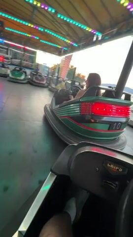The video shows a first-person view of someone riding in a bumper car at an amusement park. The bumper car is green with red accents. The person driving is wearing a grey sock and has a bracelet on their wrist.

In the background, other bumper cars can be seen, some with people inside. One bumper car in the foreground has a woman and a child in it. The amusement park appears to be outdoors, with a yellow tent-like structure overhead adorned with green lights. There are neon signs in the background, one of which appears to say "Bumper Cars." The setting appears to be daytime, possibly late afternoon or early evening, given the lighting. The overall mood seems fun and energetic, typical of a carnival or fair.