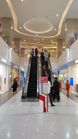This video shows a vertical view of a shopping mall interior. In the center, two escalators are visible, with several people using them. The escalator on the left is moving upwards, and the one on the right is moving downwards. A red and white signpost is placed between the two escalators, with text on it. On the left side of the mall, there are shops, including one labeled "MYSTERY." On the right side, more shops are visible, including one with a "HUAWEI" sign. Several people are walking around in the mall. The environment appears clean and well-lit, with modern architecture, including curved ceiling elements and recessed lighting. The floor is tiled.