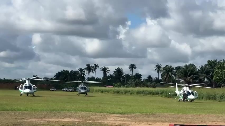 The video shows three helicopters parked on a grassy field. The helicopters are painted white with green accents. Behind the helicopters are palm trees, lush vegetation, and a few vehicles. The sky is overcast with thick, gray clouds, and patches of blue sky are visible. The rotor blades on the helicopters are still. The scene appears to be an outdoor helipad in a tropical or subtropical location.