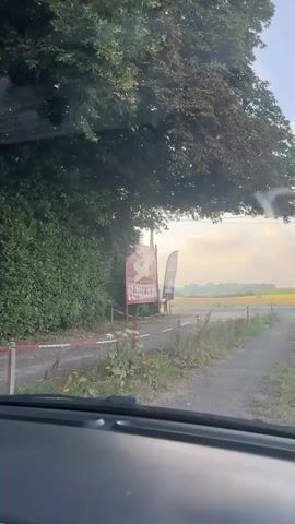 The video shows a view from inside a car looking out at a roadside scene. A large, burgundy banner with a white silhouette of a rabbit and the text "LE MANOIR DE FUMECHON" is visible, supported by two poles. To the right of the banner, a tall, black feather flag with a white symbol is planted. Behind these, a telephone pole stands. The car is on a gravel road, with a hedge on the left and fields visible in the distance. The sky is bright with some clouds. The car appears to be moving forward, and the driver's voice says "Sla links af" which translates to "Turn left."