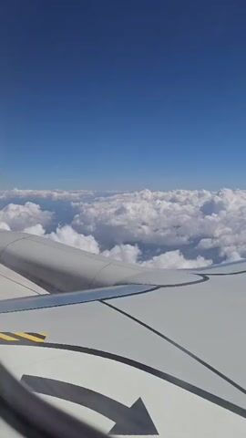 A view from an airplane window shows the sky and clouds from above. The wing of the plane is visible in the foreground, with an arrow marking the wing. The sky is clear and blue, with puffy white clouds below. The plane appears to be in flight.