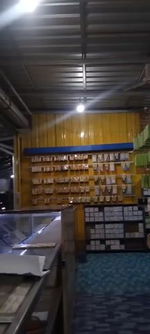 The video shows the interior of a retail store, likely selling mobile phone accessories. The walls are painted yellow and decorated with rows of small packaged items, possibly phone cases or screen protectors. Shelves are filled with more products, including boxes and hanging items. A ceiling fan is visible, and the lighting appears to be artificial, with a bright bulb illuminating the center of the store. The floor is covered in a patterned tile or carpet.