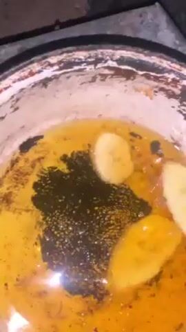 A person is deep frying sliced bananas in a weathered pot filled with yellow oil. The person carefully places the banana slices into the hot oil, where they begin to fry. The pot is heavily used, with a dark, possibly burned, residue at the bottom and along the sides. The cooking takes place under dim lighting.