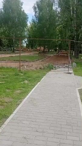 A static, eye-level shot reveals a fenced-off construction area in a park. A grey brick pathway leads towards the fenced area. The fence is made of metal posts and wire mesh, and it surrounds an area of dirt and partial construction. Behind the fence, a person is seen digging in the dirt with a shovel. The park has green grass on either side of the pathway and several tall green trees are visible in the background. The sky above is partly cloudy.