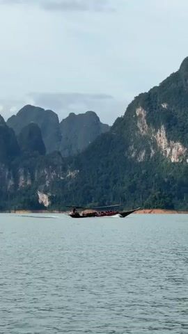 A long, narrow boat with a covered seating area and a canopy is moving across a large body of water. The boat is carrying several passengers. The background features a dramatic landscape of steep, tree-covered mountains. The sky is overcast. The water is calm with slight ripples. The boat is creating a wake as it moves to the left of the frame. The scene appears to be a scenic tourist location.