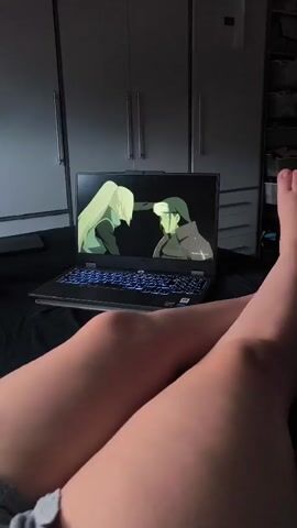 A person's legs are visible in the foreground, with their feet at the top right of the frame. They are lying down, possibly on a bed, with their legs crossed. In the background, a laptop is open and displaying an anime scene. The anime appears to be a dialogue scene with animated characters. The environment looks like a bedroom with a wardrobe in the background. The lighting is dim, suggesting it might be evening or indoors with artificial light.