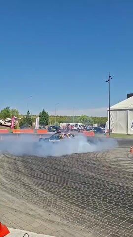 A black and yellow drifting car kicks up a large cloud of white smoke as it spins around an orange barrel barrier in a paved area. The car is angled sharply to the right, with its rear tires sliding. Several spectators are visible in the background, along with tents and other vehicles. The sky is clear and blue. An orange and white traffic cone is in the foreground on the left.