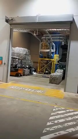 The video shows a warehouse or industrial setting. In the foreground, there's an open roll-up door with "KEEP CLEAR" painted on the floor in yellow, with gray striping underneath. Inside the warehouse, large stacks of compressed recycled materials, possibly paper, are visible. A forklift is parked near the stacks. There's also a yellow scaffolding-like structure with blue industrial machinery and pipes on top. Other equipment and containers are scattered around the floor. The lighting appears to be standard warehouse lighting.