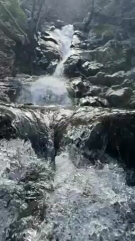 The video shows a waterfall cascading down a rocky slope. The water is white and foamy as it tumbles over the rocks. The surrounding environment appears to be a forest, with trees visible on the edges of the frame. The light is bright, creating reflections on the water's surface. The camera angle is low, providing a ground-level perspective of the waterfall.