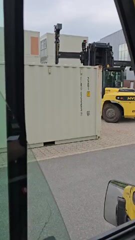 This media file captures a scene in Haren (Ems), Germany, likely in an industrial or logistics yard. The primary object of focus is a large, yellow Hyster forklift equipped with a specialized container spreader attachment. This attachment is positioned above a white shipping container, suggesting the forklift is either about to lift or has just finished lifting the container. The forklift itself is robust, with large tires suitable for uneven terrain.

In the background, several modular or prefabricated buildings, also white, are visible. These structures, along with the shipping container and the forklift, create an environment associated with warehousing, manufacturing, or shipping operations. The ground is paved with light-colored interlocking blocks, and a section of asphalt road is also present.

The weather appears overcast, contributing to a diffused, natural light. The time of day is not precisely discernible, but the lighting suggests it is daytime, possibly late morning or afternoon.

No people are clearly visible within the frame, though a figure might be inside the forklift cabin. The perspective seems to be from inside another vehicle, possibly a truck or another forklift, looking out through a window. A side-view mirror from this interior vehicle is partially visible in the foreground, reflecting a portion of the yellow forklift.

Visible text on the white shipping container includes "ELAU" and a series of numbers and letters: "22G1 ELAL U 075353 8 22G1". There is also a yellow warning label with a black symbol. On the forklift, the brand name "HYSTER" is prominently displayed on the front.

The overall impression is one of a routine operation in a busy industrial setting, where heavy machinery is used to move large containers. The scene is functional and devoid of overt emotional cues, focusing on the mechanical activity.