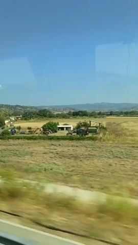 The video shows a view from a moving vehicle, likely a train, looking out at a rural landscape. The scene is dominated by a clear blue sky and rolling hills in the distance. In the foreground, there are fields of dry grass and some scattered trees. There are also a few buildings and structures, including what appear to be houses and farm buildings, along with some agricultural machinery. A road is visible in the middle ground, with a few cars on it. The perspective suggests the viewer is traveling at a moderate speed, causing a slight blur to the scenery. The overall impression is of a sunny day in the countryside.