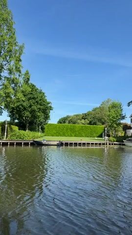 The video shows a body of water, likely a lake or wide river, under a clear blue sky. The camera appears to be on a boat moving through the water, as evidenced by the ripples and movement. Along the bank are large, well-maintained houses with docks, lush green lawns, and trimmed hedges. Several trees are visible, including a large weeping willow. One boat is docked, and a second is visible in the frame for a short time. The overall setting is affluent and well-kept. There is a person speaking, but they are off camera.
