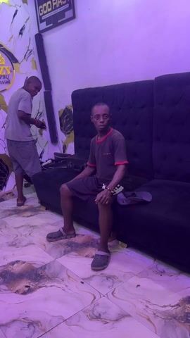 A close-up video shows a man in a brown shirt with red trim and black shorts, dancing and smiling. The man is fair-skinned and has a thin build. He stands up from a black tufted sofa. In the background, another younger man in a gray shirt and shorts is visible, standing and slightly smiling. The room has a modern interior with a marble-patterned floor, and the walls feature a design with lightning-like gold cracks against a white and black background. A sign for a barbershop "DANZ EMPIRE" can be seen on the wall. The video has a purple/blue lighting effect. The man dancing moves toward the camera, engaging the viewer with his smile.