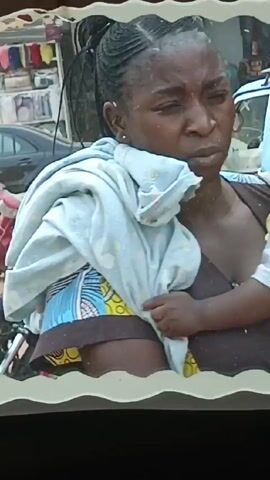 A woman wearing a blue and white patterned cloth is holding a child as she sits in a vehicle. The vehicle is yellow and there is a view of a street outside.