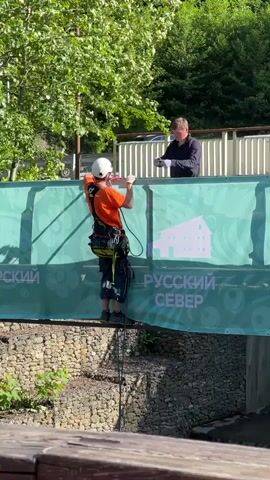 In Эстосадок, Russia, an outdoor scene unfolds under bright daylight. A man wearing a white helmet, an orange t-shirt, dark pants, and a safety harness is positioned on a narrow beam or ledge, holding onto a bar or cable above him. He appears to be preparing for a controlled descent or performing maintenance on the structure, with safety ropes attached to his harness extending downwards. Another man in a dark shirt stands on a higher platform behind a vibrant green banner, observing the activity. The banner prominently features white text, including "РУССКИЙ СЕВЕР" (Russian North) and a partial "РСКИЙ", along with a white house icon. Lush green trees form the background, and a stone-faced embankment is visible below the banner and the man in the harness.