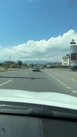 This video captures a view from a moving vehicle on a road, likely in or near Proshyan, Armenia. The scene is dominated by a wide, paved road with lane markings. Several vehicles are visible on the road, including cars and trucks, suggesting moderate traffic. In the foreground, a silver sedan is prominent, followed by other cars and a light-colored SUV. To the right of the road, there are some market stalls or vendors with tents, and merchandise, possibly produce, is displayed in bags and on a white van.

The background reveals a town or village with houses and buildings, and beyond that, a majestic mountain range capped with snow. The sky is partly cloudy, indicating daytime with a mix of sun and clouds. The overall atmosphere appears to be a typical day of travel or commerce along a scenic route.

Visible text includes license plates on the vehicles. The silver sedan has a license plate reading "37 UB 004 AM". The SUV has a license plate reading "34 TN 608 AM". There is also a sign with text partially visible in the background, reading "STUMBRAS". The presence of snow-capped mountains suggests a cooler climate or a higher elevation, and given the location context, this is likely the Aragats mountain range in Armenia. The presence of market stalls indicates a local activity, possibly a roadside market where goods are being sold. The time of day appears to be daytime, given the lighting conditions and the shadows.