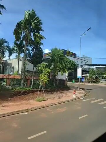The video shows a street scene with a hotel and a coffee shop. A person on a motorcycle is riding through a crosswalk. A white car is also visible in the foreground. The sky is blue and there are some trees and palm trees in the scene. The hotel has a sign that says "PHƯỚC HÙNG HOTEL" and "PHƯỚC HÙNG COFFEE TEA". There are some people walking in the background near the coffee shop. The video is filmed from a moving vehicle.
