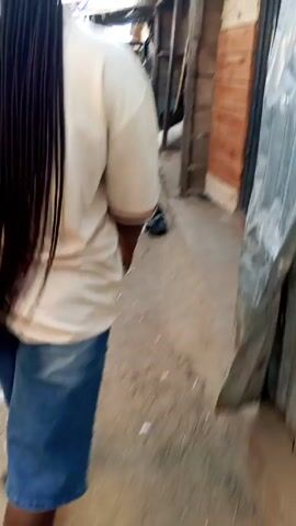 A person with long braids, wearing a white short-sleeved shirt and blue denim shorts, walks on a narrow paved area. They are holding a phone in their left hand and wearing brown flip-flops. The person walks past a parked motorcycle and other people are visible in the background. The environment appears to be a busy outdoor setting with partial shelter overhead, possibly a marketplace or similar commercial area.