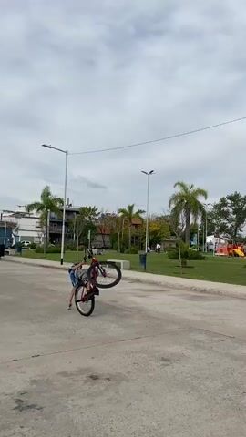 A young person wearing a helmet, blue shirt and patterned shorts, is performing a balancing act on a red bicycle on an asphalt road. The person is bent backward, supporting their weight with one hand on the ground while balancing the bike upright. The backdrop shows a park with trees, grass, benches, and a few people in the distance. In the latter half of the video, three more individuals on bicycles appear, watching the person. The sky is overcast, and there are street lights and utility poles visible.