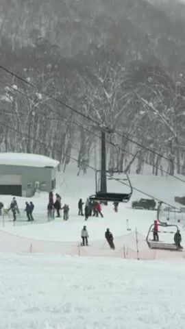 The video shows a snowy ski area on a cloudy day. A ski lift runs up the slope in the middle of the frame, with empty chairs visible. People dressed in winter clothing are scattered on the snowy ground near the base of the ski lift, some on skis or snowboards, others standing or walking. A small light-colored building is visible to the left of the ski lift, and a vehicle is parked to the right. The background consists of snow-covered trees on a hillside. It is snowing lightly.