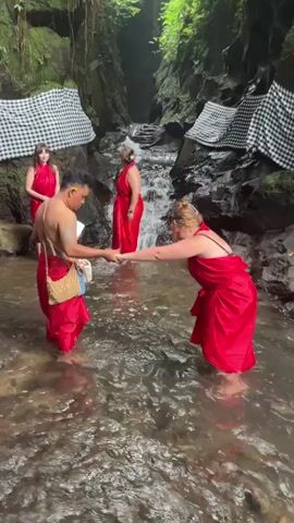 A group of four people are standing in a shallow, rocky stream in a lush, green gorge. The stream has a small waterfall in the background. Two of the people are women wearing red dresses, and two are a man and a woman also wearing red sarongs. The man in the sarong is holding hands with the woman in the sarong, and they appear to be participating in a ritual. Two other women in red dresses stand to the side, observing. White and black checkered cloths are draped on the rocks on either side of the gorge.