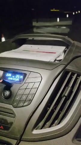 The video is a first-person view from inside a vehicle at night. The dashboard is visible, with gauges and a digital display. Papers are on the dashboard, and there's a vent. The road ahead is dark with streetlights and the headlights of other vehicles. The vehicle appears to be in motion.