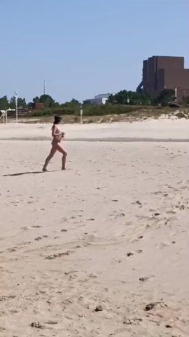 A wide shot of a sandy beach under a clear blue sky. In the foreground, the sand is wet and appears to have been recently exposed to water, forming a shallow pool. Further back, a person in a bikini walks from right to left across the sand. In the background, other beachgoers are visible, some under umbrellas, and a yellow lifeguard stand can be seen. Trees and buildings line the distant horizon. A second shot shows two children, a girl in a bikini and a boy in blue swim trunks, walking on the sand. The girl appears to be looking down. The third shot shows the same beach scene as the first, with the person in the bikini still walking in the same direction. The fourth shot shows a different part of the beach, with several people walking near a small body of water. The fifth shot is a wider view of the beach, showing the water's edge and several distant figures.