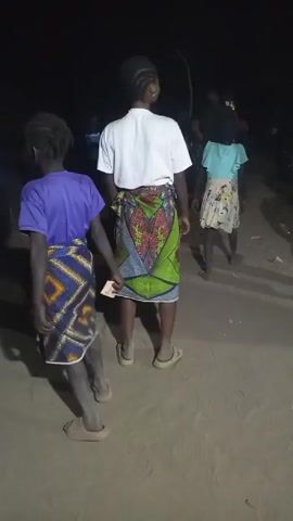 A night-time video shows a group of people dancing. The light is poor and visibility is low. The people are mostly children and women. They are wearing colorful traditional clothing. There is a sound track playing. The ground is sandy. The location is in an open area.
