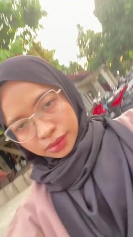 This media file is a selfie video recorded by two young women in Air Molek, Indonesia. The first woman, who is recording the video, is wearing a dark grey hijab and a light brown top. She has on large, silver-rimmed glasses and is looking directly at the camera with a neutral expression. The second woman, seated next to her, is wearing a black hijab and a red top. She is looking at the camera with a slightly confused or pouting expression.

The setting appears to be outdoors, possibly a park or recreational area, with a concrete ground visible. In the background, there are blurred images of buildings, motorcycles, and other people, suggesting a public gathering or outdoor space. The time of day seems to be daylight, but the exact time is not clear. The weather appears to be fair. The overall mood of the video is casual and informal, typical of a selfie shared among friends. There is no visible text in the media file.
