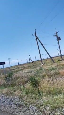 The video shows a sunny day with a clear blue sky. A car is driving along a road with a white line on the side. On the right side of the road, there is a field with green and brown grass and some wildflowers. In the distance, there are several tall metal structures, possibly power line towers, stretching across the landscape. The camera is positioned inside the car, looking out the window, and captures the passing scenery.