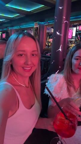 Three women are enjoying cocktails at a crowded bar. They are all smiling and appear to be having a good time. The bar is brightly lit with pink and blue lights. There are other people in the background, and the bar appears to be busy. One of the women is wearing a white tank top with a gold chain, another is wearing a white, fluffy shirt, and the third is wearing a white long-sleeve shirt with a gold chain. They are all holding their drinks up in a toast.